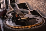 A glossy, black acoustic guitar rests in an open, plush-lined case. The guitar features a traditional tear-drop-shaped soundhole with elegant detailing around the edges. Its strings stretch tautly across the body, and the ambient lighting casts soft reflections on its polished surface.