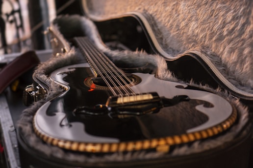 A glossy, black acoustic guitar rests in an open, plush-lined case. The guitar features a traditional tear-drop-shaped soundhole with elegant detailing around the edges. Its strings stretch tautly across the body, and the ambient lighting casts soft reflections on its polished surface.