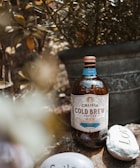 A bottle of Californian cold brew coffee is positioned outdoors on a rock surface. The bottle is brown with a decorative label indicating the coffee is mocha flavored and concentrated. Surrounding the bottle are stones with engraved words like 'love.' There's a large decorative plant pot in the background along with green foliage, giving an earthy ambiance.