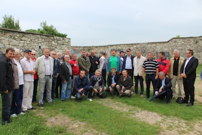 A group of men gathered outdoors, standing and kneeling on grass with a stone wall in the background. The men are dressed in casual and semi-formal attire, indicating a social or community event. The weather appears mild, with overcast skies.