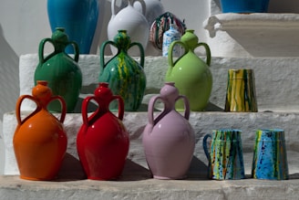 A collection of large, colorful ceramic jugs and cups arranged on stepped shelves. The jugs are in various colors including green, orange, red, and purple, each with a unique glossy finish. The cups have intricate, multicolored patterns. The setting is outdoors, with bright sunlight casting shadows.