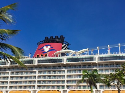 A large cruise ship with multiple decks and balconies is seen under a clear blue sky. The ship features a prominent red funnel with a cartoon character logo. Palm leaves are visible in the foreground, adding to the tropical feel of the scene.