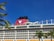 A large cruise ship with multiple decks and balconies is seen under a clear blue sky. The ship features a prominent red funnel with a cartoon character logo. Palm leaves are visible in the foreground, adding to the tropical feel of the scene.
