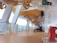 A deck on a cruise ship with wooden flooring and white structural beams overhead. Several life boats are suspended above. There are a few deck chairs lined up against the wall and a sign indicating a muster station. The area appears clean and organized, with ambient natural light.