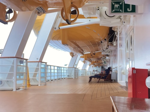 A deck on a cruise ship with wooden flooring and white structural beams overhead. Several life boats are suspended above. There are a few deck chairs lined up against the wall and a sign indicating a muster station. The area appears clean and organized, with ambient natural light.