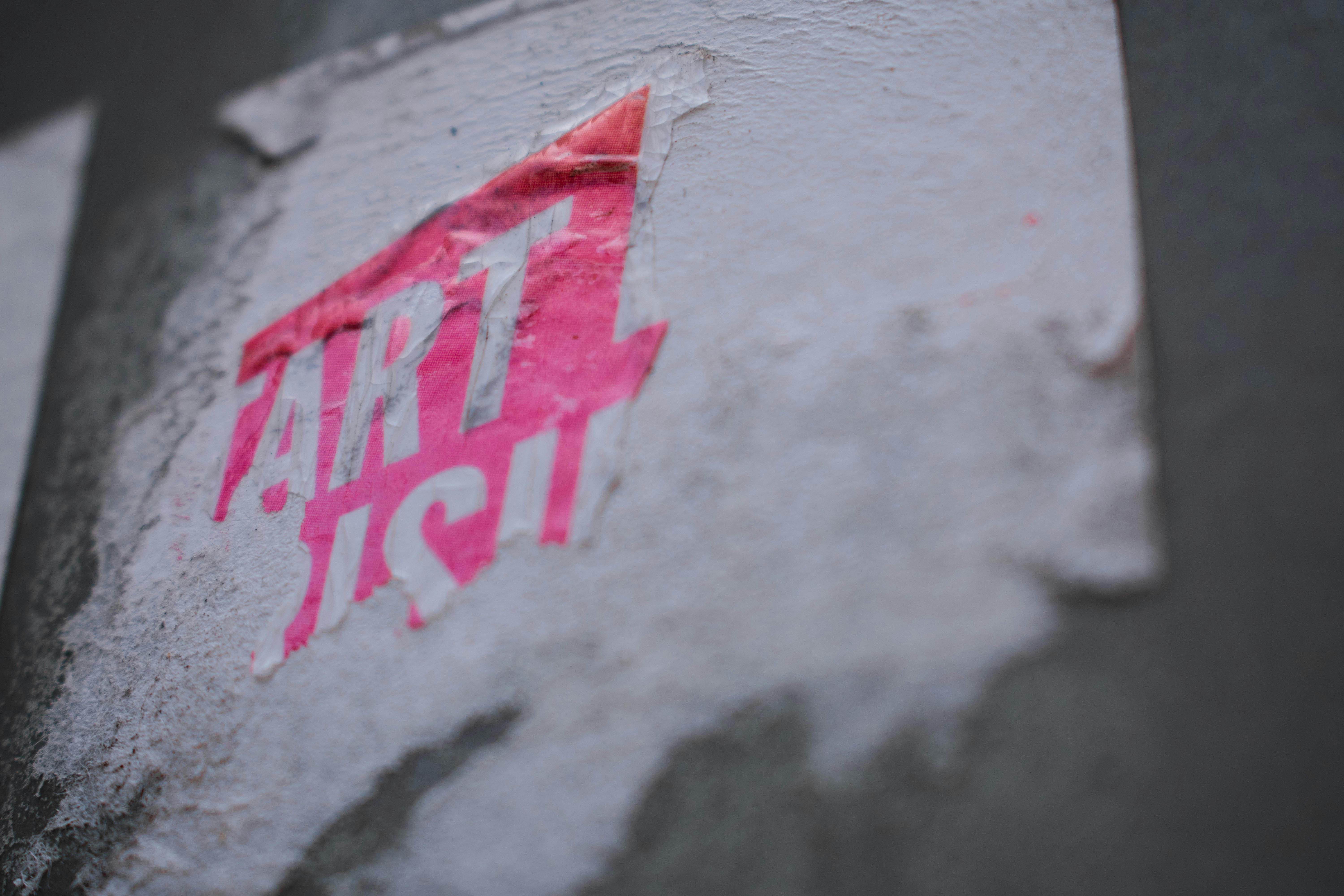 A partially torn and weathered sticker with the word 'ART' prominently visible in bright pink and partially obscured white lettering, adhered to a textured, off-white surface.