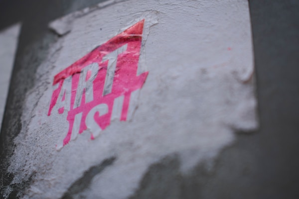 A partially torn and weathered sticker with the word 'ART' prominently visible in bright pink and partially obscured white lettering, adhered to a textured, off-white surface.