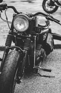 A close-up shot of a vintage-style motorcycle captured in black and white. The focus is on the front part, showcasing the round headlight, handlebars, and front tire. The texture of the tire and the metallic components are prominent. Another motorcycle is partially visible in the background.