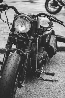 A close-up shot of a vintage-style motorcycle captured in black and white. The focus is on the front part, showcasing the round headlight, handlebars, and front tire. The texture of the tire and the metallic components are prominent. Another motorcycle is partially visible in the background.