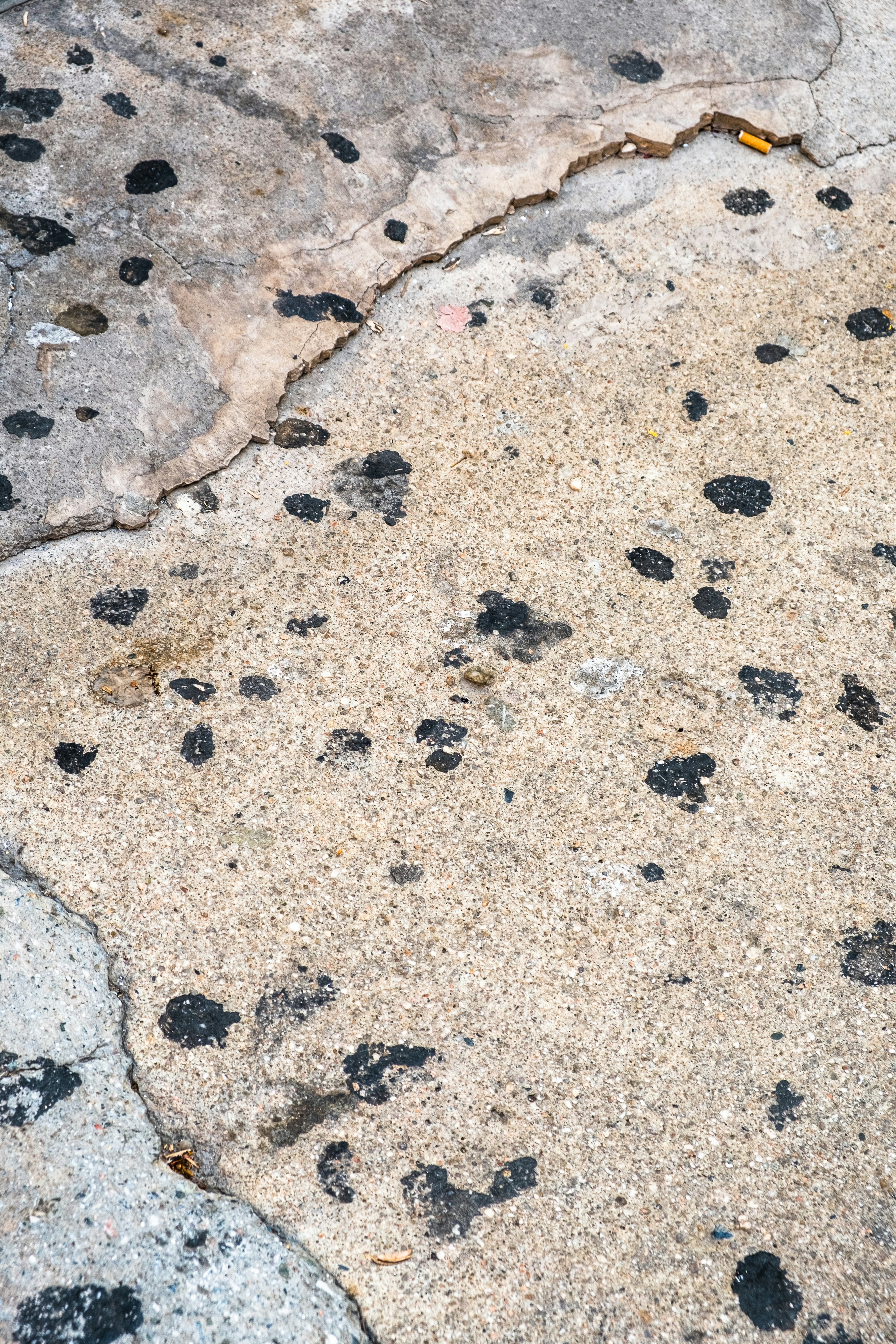 Cracked and dirty concrete sidewalk with dark black stains, oil marks, and years of urban grime, dim overcast lighting