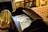 An old wooden desk with a raised backrest holds a stack of vintage newspapers. A multicolored, patterned bedspread can be seen to the right, and there is a chair draped with a piece of fabric positioned on the left. A bookshelf filled with books is visible in the background.