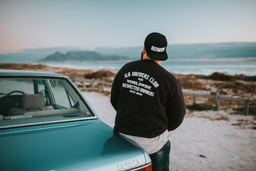 A person wearing a black cap and a dark jacket with the words 'LB Drivers Club Worldwide Respected Owners' stands leaning against a vintage car near a coastal landscape. The ocean and a mountainous horizon are visible in the background under a cloudy sky.
