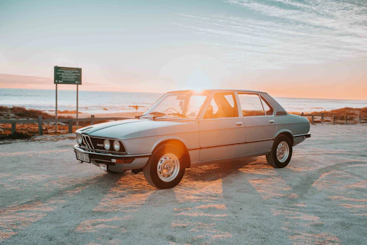 A gleaming vintage car parked on a winding coastal road at sunset, with the sea shimmering in the background.