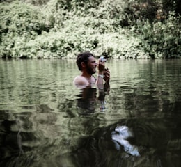A man is standing waist-deep in a body of water surrounded by dense green foliage. He is holding a camera up to his face as if taking a photograph. The water creates a reflective surface, capturing the greenery and adding a tranquil feel to the scene.