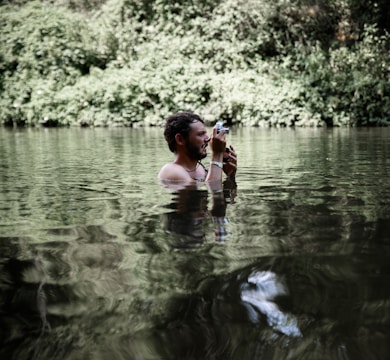 A man is standing waist-deep in a body of water surrounded by dense green foliage. He is holding a camera up to his face as if taking a photograph. The water creates a reflective surface, capturing the greenery and adding a tranquil feel to the scene.
