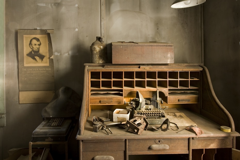 An old wooden desk with cubbyholes filled with papers and small items. A vintage typewriter sits in the center of the desk, surrounded by various tools, a pair of goggles, and stacks of paper. On the wall, a sepia-toned poster of a historical figure is visible, along with an aged jug and a nearby hat, suggesting a setting from the early 20th century. The workspace exudes a sense of antiquity and nostalgia.