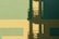 A minimalist composition featuring geometric shapes in shades of green and yellow. The interplay of light and shadow creates a complex pattern on a flat surface. A shadow resembling a structure with railings and vertical lines is cast over the blocks of color.