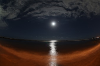 A serene night scene featuring a bright, full moon illuminating a calm ocean. The moonlight creates a shimmering pathway across the water's surface, framed by a cloudy night sky. The horizon is faintly visible, and the overall atmosphere is peaceful and contemplative.
