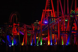 Brightly colored amusement park roller coaster features prominently with vivid red, blue, green, and purple lights illuminating the tracks and structure. The scene is set at night, creating a dramatic and vibrant atmosphere with the lights casting dynamic shadows.