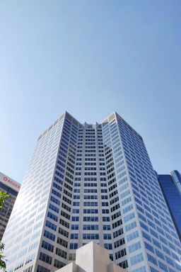 A modern skyscraper with a reflective glass facade towers into a clear blue sky. The building's geometric design features numerous windows, creating a grid-like pattern across its surface. Other high-rise buildings are partially visible in the background.