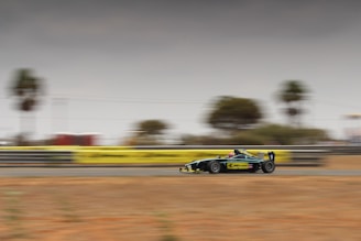 A formula race car is moving at high speed on a racetrack. The background is blurred, emphasizing the motion and speed. The car has a sleek design with a dark color and yellow accents. Trees and a barrier line the track, and the sky is overcast.