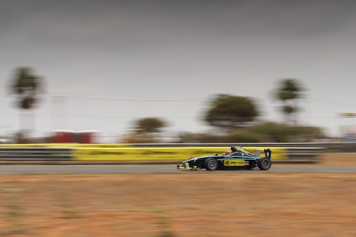 A formula race car is moving at high speed on a racetrack. The background is blurred, emphasizing the motion and speed. The car has a sleek design with a dark color and yellow accents. Trees and a barrier line the track, and the sky is overcast.