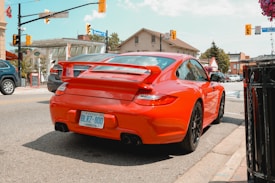 A bright red sports car is parked on the street at an intersection in a small town. The license plate indicates it is from Ontario. Surrounding the car are other vehicles, buildings with signage, and traffic lights. The scene has clear skies and a casual urban atmosphere.