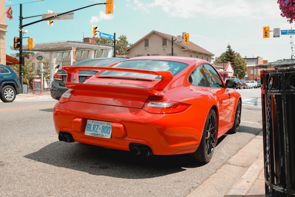 A bright red sports car is parked on the street at an intersection in a small town. The license plate indicates it is from Ontario. Surrounding the car are other vehicles, buildings with signage, and traffic lights. The scene has clear skies and a casual urban atmosphere.