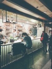 A dimly lit bar with several people sitting and standing around the counter. The bar counter is adorned with various bottles and items, and the background features exposed brick walls. Metal shelves hang from the ceiling, holding bottles and glasses. The atmosphere is casual and relaxed, with a few patrons engaging in conversation.