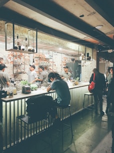 A dimly lit bar with several people sitting and standing around the counter. The bar counter is adorned with various bottles and items, and the background features exposed brick walls. Metal shelves hang from the ceiling, holding bottles and glasses. The atmosphere is casual and relaxed, with a few patrons engaging in conversation.