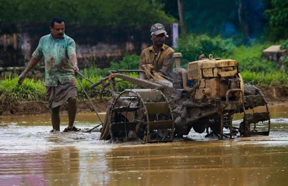 Two men navigate a muddy field with a plowing machine, which is coated in wet soil. The scene is set in a rural area with greenery in the background, and both men and machine are engaged in agricultural work.