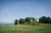 A solitary house sits atop a gently sloping hill, surrounded by a cluster of lush green trees under a vast clear blue sky. In the distance, a paraglider with a colorful parachute gracefully glides through the air, emphasizing the openness and tranquility of the landscape.