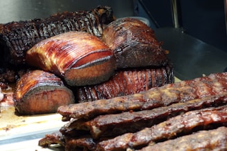Slices of roasted meat, possibly beef, arranged on a wooden cutting board. The meat appears juicy with a deep brown, caramelized crust and a tender red interior. In the foreground, there are ribs with a similar glazed finish, and some charred edges are visible.