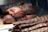 Slices of roasted meat, possibly beef, arranged on a wooden cutting board. The meat appears juicy with a deep brown, caramelized crust and a tender red interior. In the foreground, there are ribs with a similar glazed finish, and some charred edges are visible.