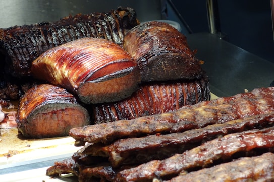 Slices of roasted meat, possibly beef, arranged on a wooden cutting board. The meat appears juicy with a deep brown, caramelized crust and a tender red interior. In the foreground, there are ribs with a similar glazed finish, and some charred edges are visible.