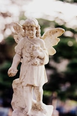 A weathered stone statue of an angelic figure with wings, holding a small animal in its arms. The angel is depicted with curly hair and a serene expression, standing on a pedestal with a muted, blurred background of greenery and soft, natural light.