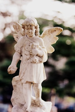 A weathered stone statue of an angelic figure with wings, holding a small animal in its arms. The angel is depicted with curly hair and a serene expression, standing on a pedestal with a muted, blurred background of greenery and soft, natural light.