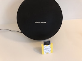 A large black speaker with the brand name 'harman/kardon' is placed on a light-colored surface. Beside it is a small rectangular box with a yellow frame and a label that reads 'Your Tropical Vacation Soundtrack Assisted by Michel Gaubert.' A black cable is visible, leading to the speaker.