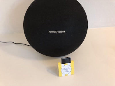 A large black speaker with the brand name 'harman/kardon' is placed on a light-colored surface. Beside it is a small rectangular box with a yellow frame and a label that reads 'Your Tropical Vacation Soundtrack Assisted by Michel Gaubert.' A black cable is visible, leading to the speaker.