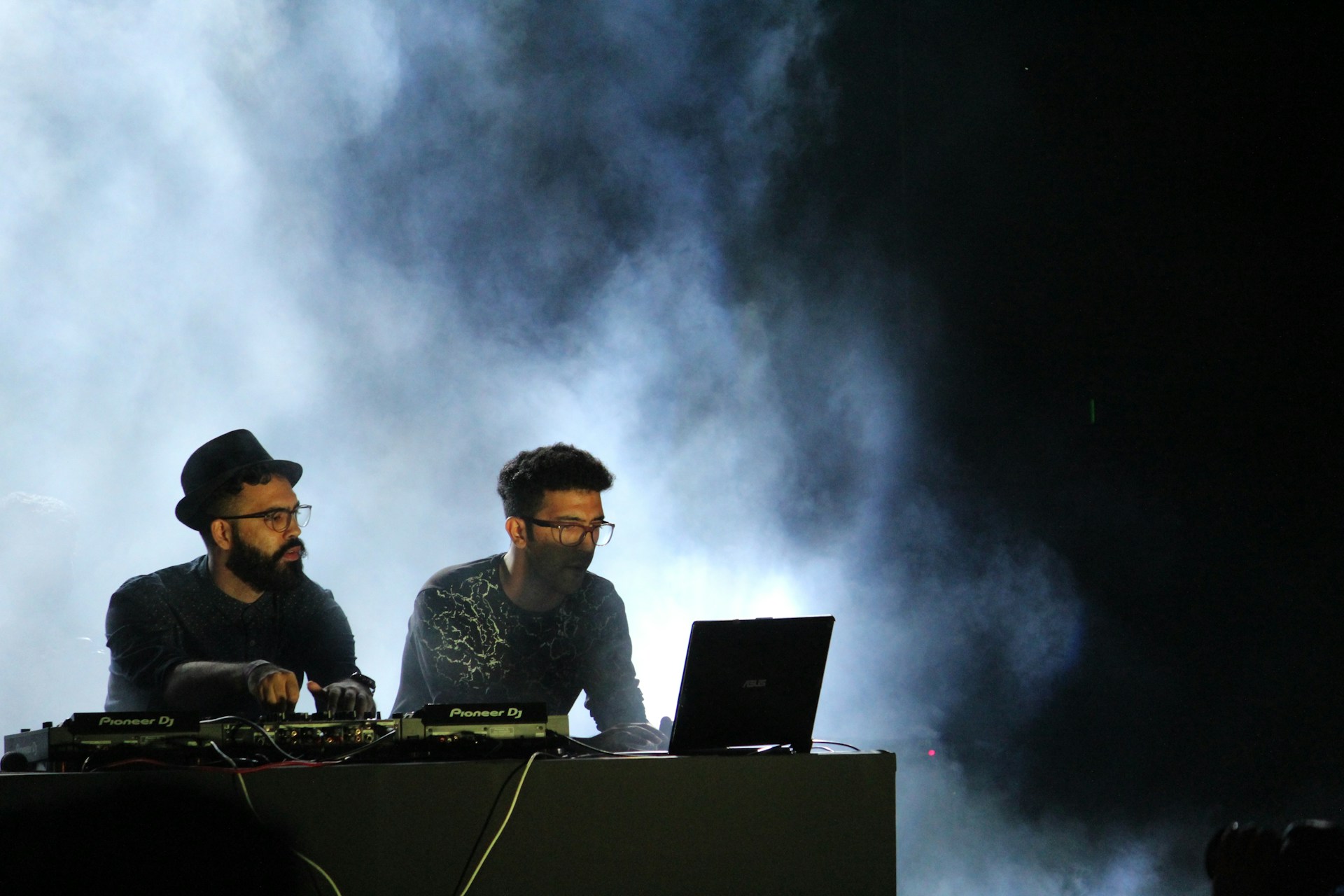 Two DJs are performing on stage, surrounded by smoke effects. One DJ is wearing a hat and glasses while the other has a beard and also wears glasses. They are focused on their equipment, which includes a laptop and mixer decks. The lighting is dim, with a blue haze from the smoke creating an atmospheric effect.