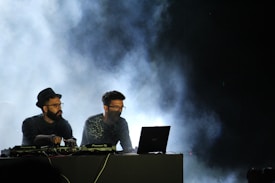 Two DJs are performing on stage, surrounded by smoke effects. One DJ is wearing a hat and glasses while the other has a beard and also wears glasses. They are focused on their equipment, which includes a laptop and mixer decks. The lighting is dim, with a blue haze from the smoke creating an atmospheric effect.