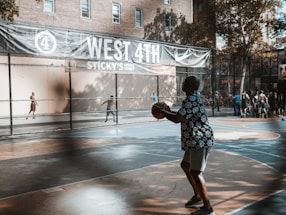 A lively outdoor basketball court with several players and people watching. One player in a patterned shirt is holding a basketball, preparing to shoot. The court has vibrant blue and orange colors. A large banner above reads 'West 4th' with references to local businesses. Trees and a building can be seen in the background, casting shadows on the court.