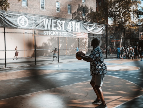 A lively outdoor basketball court with several players and people watching. One player in a patterned shirt is holding a basketball, preparing to shoot. The court has vibrant blue and orange colors. A large banner above reads 'West 4th' with references to local businesses. Trees and a building can be seen in the background, casting shadows on the court.