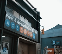 A subway entrance sign for West 4 St-Washington Square Station, displaying several subway lines including A, C, E, B, D, F, and M with a disability access symbol. There is a traffic light and nearby building signage visible in the background.