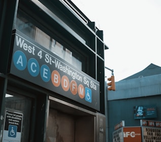 A subway entrance sign for West 4 St-Washington Square Station, displaying several subway lines including A, C, E, B, D, F, and M with a disability access symbol. There is a traffic light and nearby building signage visible in the background.