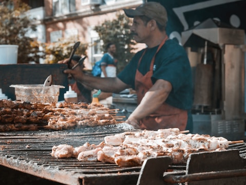 A street vendor grills various skewers of meat on a large charcoal barbecue. The vendor, wearing a blue shirt and red apron, uses tongs to handle the food. In the background, there are condiments, other kitchen equipment, and blurred figures of people and urban buildings.