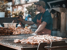 A street vendor grills various skewers of meat on a large charcoal barbecue. The vendor, wearing a blue shirt and red apron, uses tongs to handle the food. In the background, there are condiments, other kitchen equipment, and blurred figures of people and urban buildings.