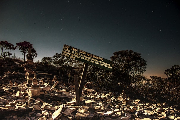 A nighttime scene with a starlit sky above a rocky area dotted with stacked stones. A wooden sign in Portuguese is prominently displayed among the rocks. The landscape has scattered trees in the background, contributing to a serene and mysterious atmosphere.