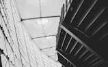 A black and white architectural photograph featuring geometric structures, including a modern roof with fabric stretched across metal beams and an angular ceiling design. The image captures the contrast between light streaming through translucent surfaces and dark shadow areas.