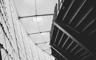 A black and white architectural photograph featuring geometric structures, including a modern roof with fabric stretched across metal beams and an angular ceiling design. The image captures the contrast between light streaming through translucent surfaces and dark shadow areas.